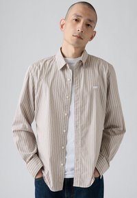 Camicia a righe beige e marrone chiaro, in tessuto di cotone, maniche lunghe, con un piccolo logo sul petto sinistro, indossata sopra una maglietta bianca.