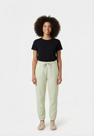 Femme aux cheveux bouclés portant un t-shirt noir à manches courtes, un pantalon de survêtement vert clair et des baskets beige, debout et souriant sur un fond blanc.