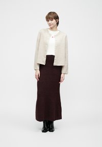 Beige wollen cardigan met ronde hals en knopen aan de voorkant, gecombineerd met een bordeauxrode gebreide maxi rok. Model draagt zwarte laarzen.