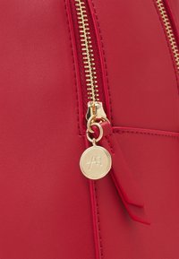 Sac en similicuir rouge avec une fermeture éclair dorée proéminente et des éléments métalliques, accompagné d'une étiquette ronde en or embossée d'initiales.