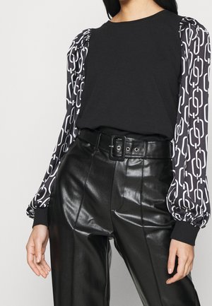Kvinde iført sort top med puffærmer i hvidt kædemønster og sorte læderbukser med høj talje og bredt bælte.