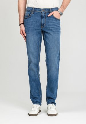 Mężczyzna w niebieskich dżinsach i białych tenisówkach, z jedną ręką w kieszeni, stojący na tle jasnego, jednolitego tła.