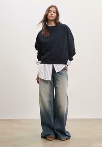 Felpa navy con scollo a costine, indossata sopra una camicia bianca a righe, abbinata a jeans in denim sbiadito a gamba larga e scarpe platform color pelle.