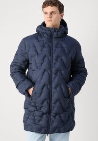 Temno modra pufasta bunda z zigzag prešitjem, kapuco, zadrgo spredaj in stranskimi žepi. Material se zdi mehak in toplotno izoliran.