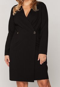 Svart omlottklänning med långa ärmar, V-ringning och en dekorativ guldfärgad knappdetalj i midjan. Tillverkad av ett mjukt, lätt tyg.