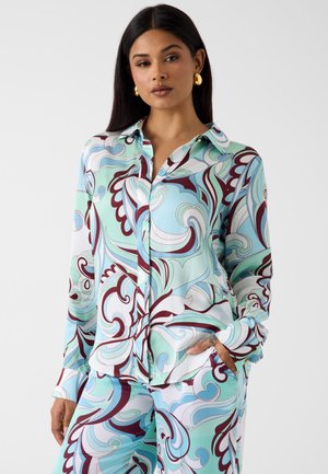 Femme aux longs cheveux foncés portant des boucles d'oreilles en or et un ensemble chemise à boutons et pantalon assortis à motifs bleu, blanc et bordeaux.
