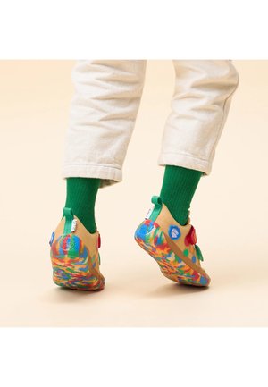 Füße in beigen Schuhen mit bunten Sohlen und grünen Socken, die auf einer beigen Fläche stehen, Hosenbeine über den Knöcheln hochgekrempelt.