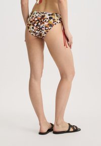 Bikini-Höschen mit Leopardenmuster aus einem glatten Stoff. Verfügt über verstellbare rote Bindungen an den Seiten. Sichtbare schwarze flache Sandalen.