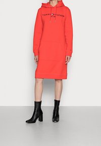 Röd huvtröjklänning med logotyp, framficka och ribbad slutning, kombinerad med svarta ankelskor med hög klossklack.