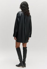 Schwarzes, oversized Hemdkleid aus Kunstleder mit langen Ärmeln, glatter Textur und hohen Seitenschlitzen, kombiniert mit kniehohen schwarzen Stiefeletten mit Absatz.