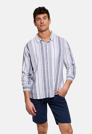 Hombre con camisa blanca de manga larga con rayas verticales grises y pantalones cortos azul marino, de pie con una mano en el bolsillo contra fondo blanco.