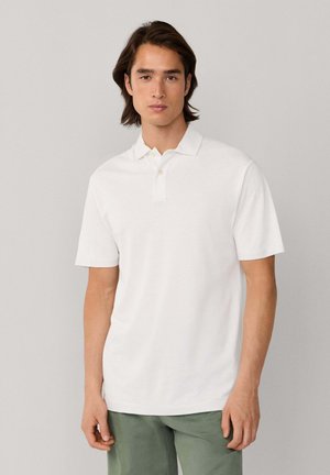 Joven con cabello castaño de longitud media, vestido con una camiseta tipo polo blanca de manga corta y pantalones verdes, sobre un fondo liso de color gris claro.