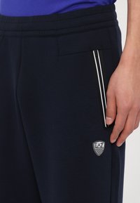 Pantaloni jogger blu navy con una texture liscia, dotati di una tasca laterale con zip e dettagli bianchi, e un patch del logo EA7 sopra la tasca.