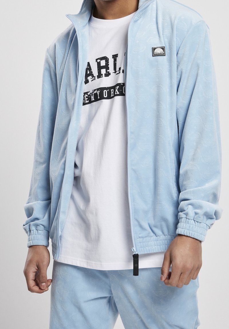 Homme portant une veste zippée à motif bleu clair et un pantalon assorti par-dessus un t-shirt blanc avec le texte noir "NEW YORK".