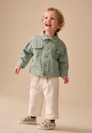 Tout-petit aux cheveux blonds bouclés souriant, portant une veste verte boutonnée, un pantalon crème et des baskets beiges, debout sur un fond beige uni.