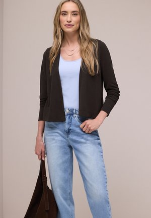 Donna bionda che indossa un cardigan nero sopra una maglia blu con jeans azzurri, tenendo una borsa marrone scuro in una mano.