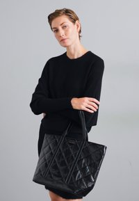 Sac fourre-tout en cuir matelassé noir avec deux poignées robustes et une base plate. Présente un motif en forme de losange et un détail de logo subtil.