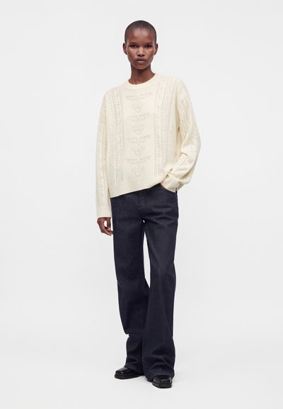 Personne portant un pull en laine crème avec des motifs de feuilles, un pantalon large foncé et des chaussures noires, debout avec une main dans la poche.