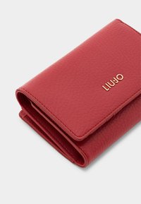 Punane tekstureeritud nahast rahakott, millel on volditud disain. Sellel on kuldne "LIU JO" logo, nähtav õmblus ja kompaktne, ristkülikuline kuju.