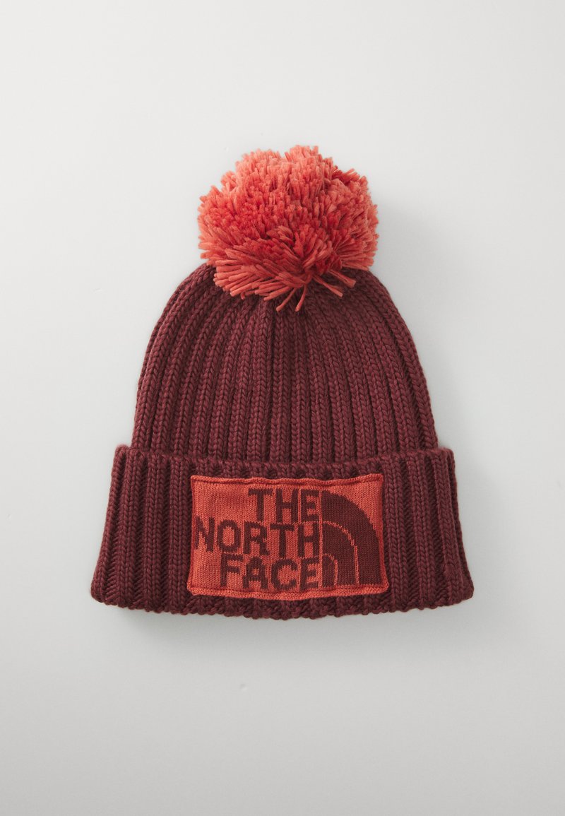 Rød strikket hue med en stor pom-pom på toppen, en foldet skygge og et rektangulært mærke med "THE NORTH FACE" logo i sort.