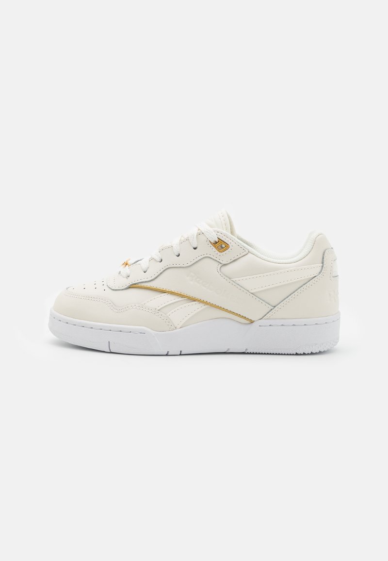 Baskets basses de couleur crème avec semelle blanche, présentant des perforations subtiles et des accents dorés sur la partie supérieure et les œillets des lacets.