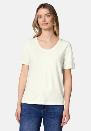 Kobieta z jasnobrązowymi włosami, ubrana w białą, gładką koszulkę z krótkim rękawem oraz niebieskie dżinsy, stojąca na tle białego tła.