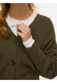 Cardigan lavorato a maglia verde oliva con scollo a V, motivi strutturati e chiusura con un solo bottone marrone, indossato sopra una camicia bianca.