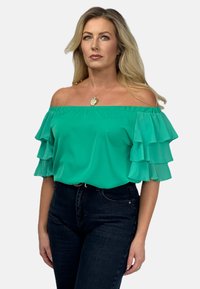 Zielona bluzka na jedno ramię z warstwowymi falbanami, wykonana z lekkiego materiału. Połączona z ciemnymi dżinsami i wisiorkiem na szyi.
