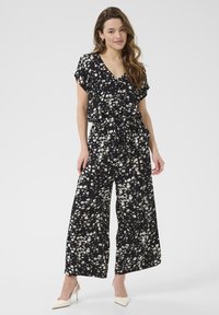 Svart blommig jumpsuit med vida ben, korta ärmar och en cinchad midjedetail. Har ett subtilt vitt blommönster genom hela.