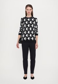 Strikket svart og hvit genser med et geometrisk trekantmønster, kombinert med svarte slim-fit bukser og spisse hæler.