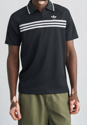 Person trägt ein schwarzes kurzärmeliges Poloshirt mit weißen Streifen und einem Logo, kombiniert mit olivgrünen Hosen, vor einfachem Hintergrund.