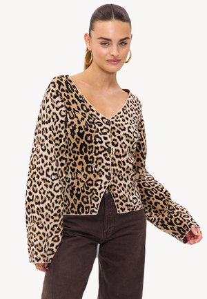 Femme aux cheveux tirés en arrière portant des boucles d'oreilles en or, un cardigan à motifs léopard à boutons et un pantalon marron foncé sur fond blanc.