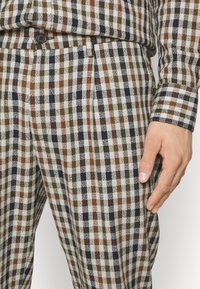 Rutiga byxor med en blandning av bruna, gröna och marinblå rutor på en vit bakgrund. Tyget har en strukturerad yta och inkluderar en framficka.