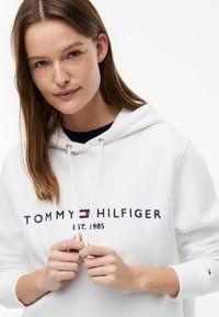 Junge Frau mit glatten braunen Haaren, die einen weißen Tommy Hilfiger Hoodie trägt und die Kordeln mit beiden Händen hält.