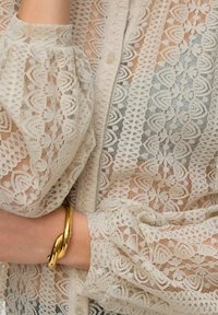 Ljust beige blus i spets med intrikata blommönster, knäppning med knappar och puffärmar. Har ett guldarmband på handleden.