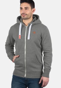 Grijze zip-up hoodie met voorzakken, witte trekkoorden en een oranje logo. Zacht materiaal met ribgebreide manchetten en zoom.