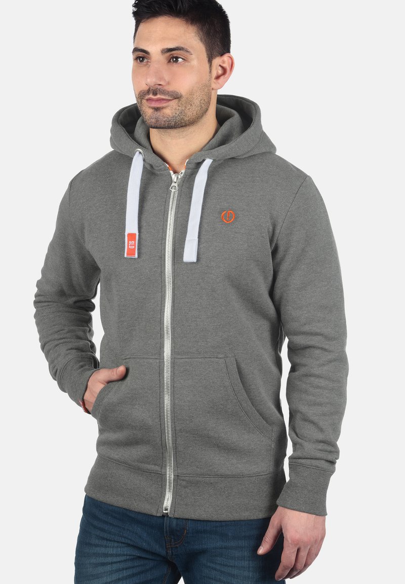 Grijze zip-up hoodie met voorzakken, witte trekkoorden en een oranje logo. Zacht materiaal met ribgebreide manchetten en zoom.