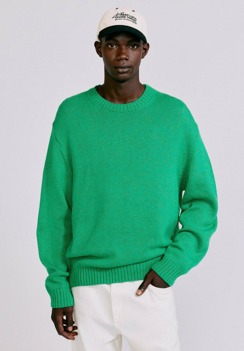 Homme portant un pull en maille vert vif, un pantalon blanc et une casquette blanche de baseball avec un texte noir, debout devant un fond clair uni.