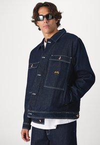 Casaco de ganga em azul escuro, com fecho de botão, duas bolsos frontais e costuras em contraste. Inclui detalhe do logótipo no bolso esquerdo.