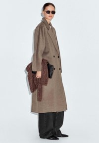 Cappotto lungo e sartoriale di colore marrone con design doppiopetto. Maglione texturizzato di un rosso scuro appoggiato su una pochette nera. Pantaloni larghi neri e scarpe nere.