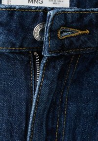 Tmavomodré džínsové nohavice s čiastočne zapnutým kovovým zipsom, kovovým gombíkom s vygravírovanou značkou a viditeľným žltým prešitím na tkanine.