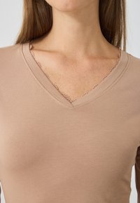Top marrone chiaro a scollo a V con pizzo sul colletto. Realizzato in materiale morbido, con texture liscia e design aderente.