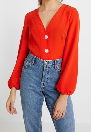 Kobieta w jasnoczerwonej bluzce z dekoltem w serek, z dwoma białymi guzikami i długimi bufkami na rękawach, w połączeniu z niebieskimi dżinsami z wysokim stanem.