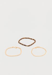 Trois bracelets sur une surface unie : un bracelet marron en perles, une chaîne en or à maillons plats, et une chaîne en or à maillons arrondis.