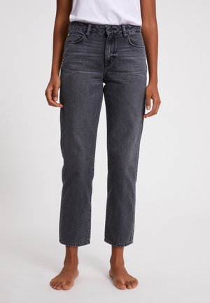 Personne portant un jean gris foncé taille haute à jambes droites et une chemise blanche, debout pieds nus sur un fond uni clair.