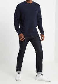 Maglione a maglia blu navy con scollo rotondo, maniche e orlo a coste, abbinato a jeans in denim scuro arrotolati ai polsi e sneaker bianche.
