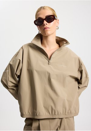Chaqueta beige con cremallera, de tipo pulóver con cuello alto y dobladillo elástico. Hecha de material ligero. Combina con gafas de sol oscuras estilo ojos de gato.