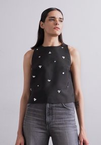 Črn brezrokavnik z ladijskim izrezom, okrašen s kovinskimi dodatki, razpršenimi po tkanini. Uporabljen skupaj z sivo obleko z visokim pasom.