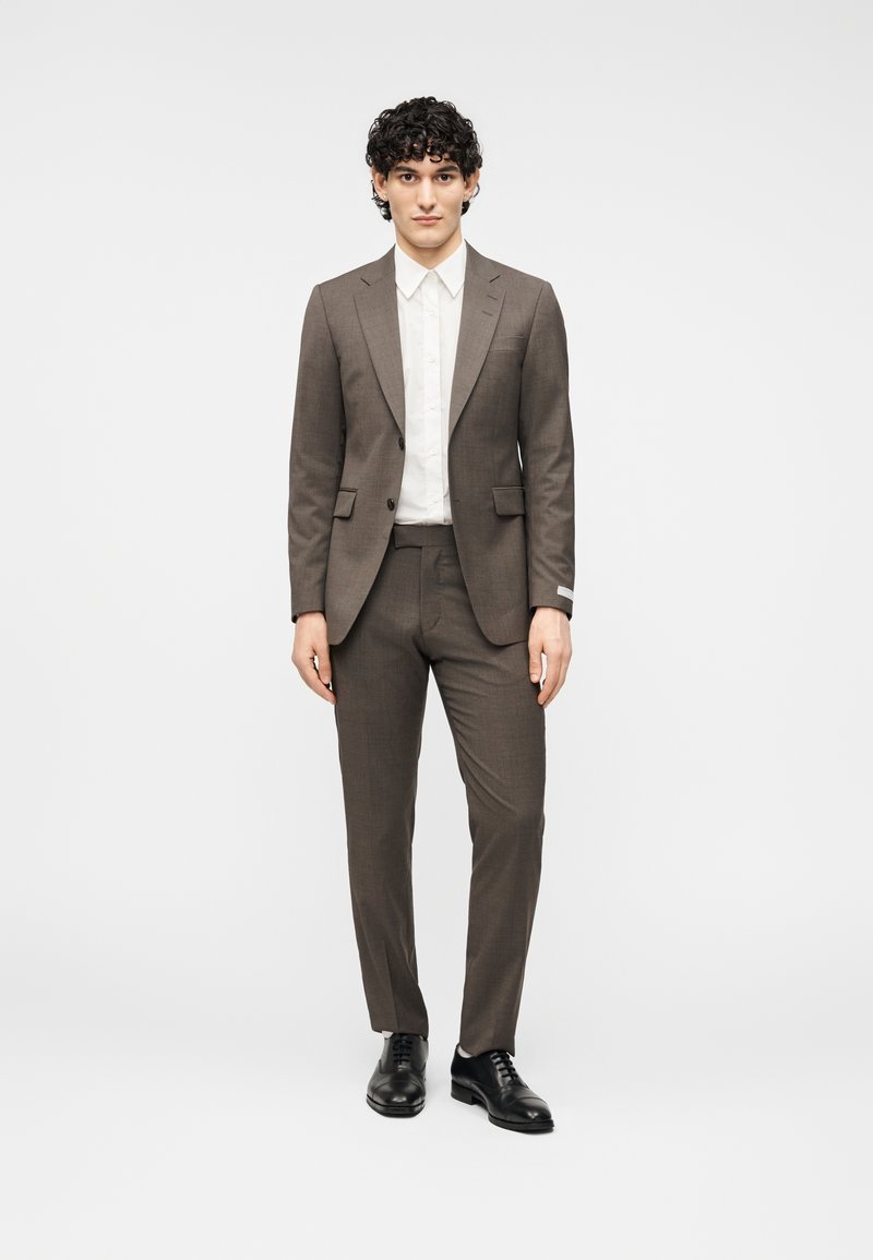 Καφέ κοστούμι με εφαρμοστή σχεδίαση, λευκό πουκάμισο από μέσα και μαύρα παπούτσια. Διαθέτει λεία υφή και αιχμηρούς γιακάδες για μια επίσημη εμφάνιση.