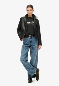 Chaqueta de moto de cuero negro, camiseta gráfica negra, jeans de mezclilla azul holgados y botas negras con cordones, con detalles de costura prominentes.